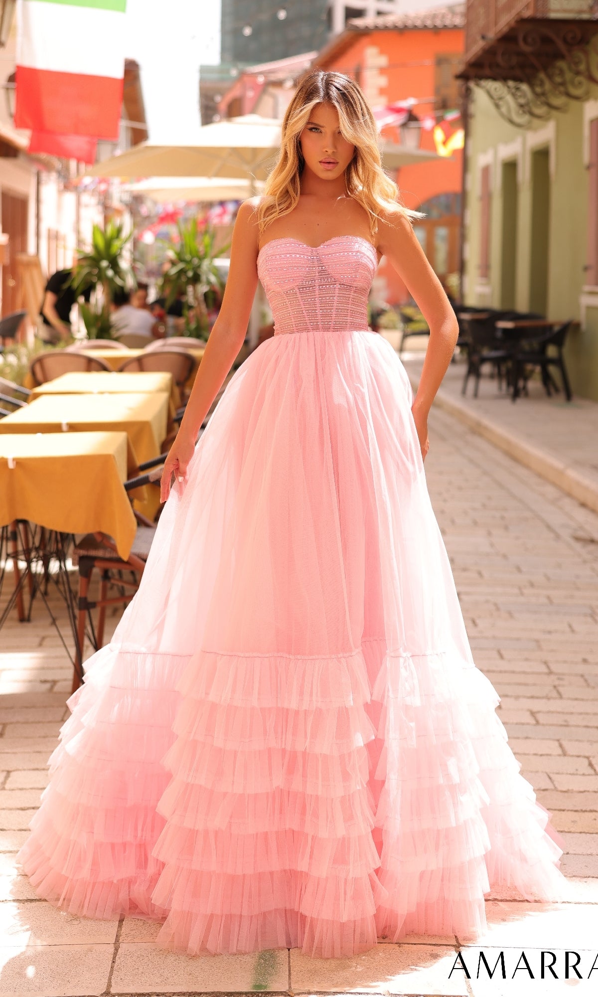 A woman stands on a cobblestone street wearing the Amarra 88277 strapless prom ball gown featuring a light pink tulle A-line ruffled skirt. Outdoor caf‚ tables are visible in the background.